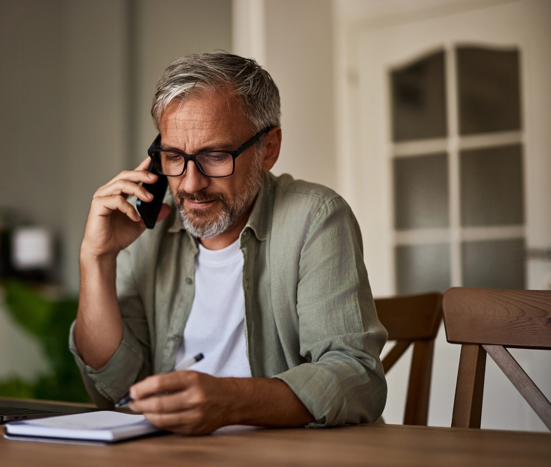 older man having a phone call and writing down notes on notepad while sitting at a wooden table.